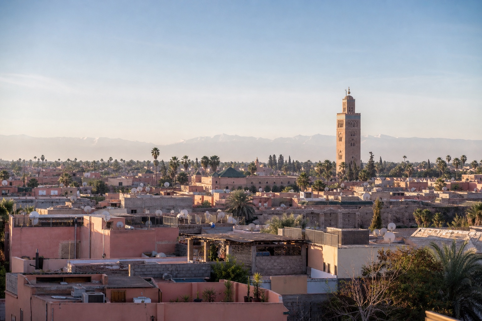 Marrakech medina rooftops, the Koutoubia minaret, palms, and the distant Atlas Mountains in morning light
