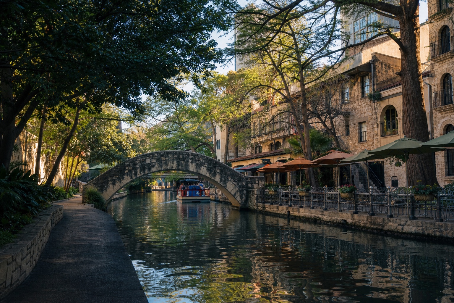 Shaded San Antonio River Walk with a stone bridge, river boat and historic riverside buildings